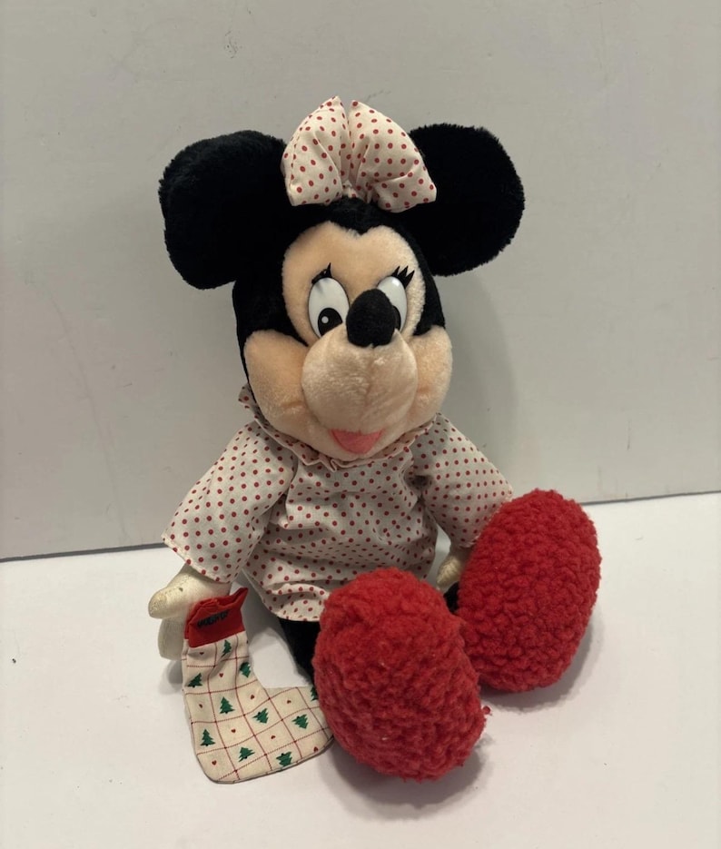 May include: A plush Minnie Mouse doll with black ears, a pink bow with red polka dots, and a pink and white polka dot dress. The doll has red fuzzy feet and a Christmas stocking.