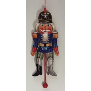 May include: A hanging nutcracker ornament featuring a black hat with a white hammer and pick emblem, a blue uniform with red accents, a white beard, and articulated legs. A red string with a wooden bead extends from the bottom for movement.