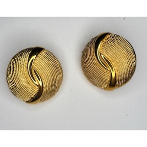 May include: A pair of round gold-tone earrings featuring a textured, cross-hatched surface pattern. Each earring is bisected by a smooth, polished, curved gold-tone band that sweeps across the center of the circular design.