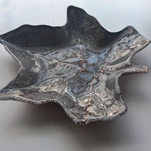 May include: A decorative leaf-shaped bowl with a textured, marbled appearance. The bowl is primarily dark gray with lighter gray and brown veining, creating a naturalistic design. The edges of the bowl are gently curved.