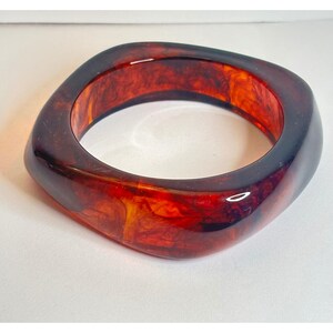 May include: A thick, square-shaped bangle bracelet featuring a polished, translucent finish with swirling patterns of deep amber, dark brown, and bright orange hues throughout the material, creating a marbled effect.