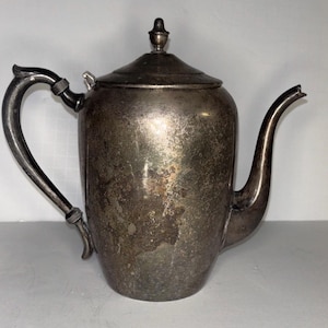 Puede incluir: Una tetera vintage de color plateado con un pico y asa curvados. La tetera tiene una tapa con un pequeño adorno y muestra signos de edad y desgaste. El diseño es clásico y elegante, adecuado para servir té o café.