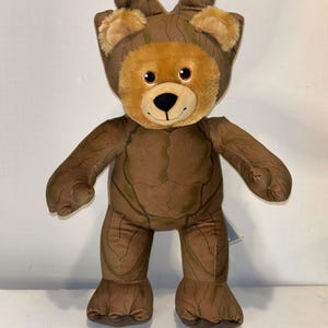 May include: A brown plush teddy bear wearing a Groot costume. The bear has a light brown face and ears, with a black nose and smiling mouth. The costume is detailed with a tree bark pattern.