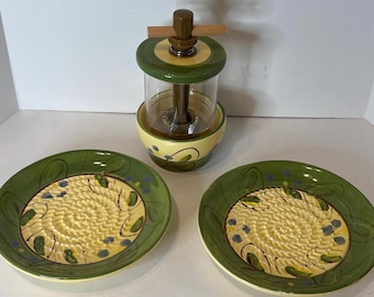 May include: A kitchen set featuring a butter churn and two ceramic plates. The churn has a glass container with a wooden handle. The plates are green with a yellow center and decorative patterns. A charming addition to any kitchen.