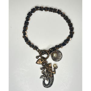 May include: A copper-toned bracelet featuring a chain of small square links and a toggle clasp. The bracelet includes three charms: a mermaid holding a small pearl, a seashell with a pearl center, and a small heart charm stamped with USA.