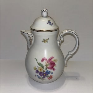 Op de afbeelding: Witte porseleinen koffiepot met deksel en handvat. De pot is versierd met een bloemenontwerp met roze, paarse, gele en blauwe bloemen. Gouden randen en handvat. Het deksel is bekroond met een roosvormige knop.