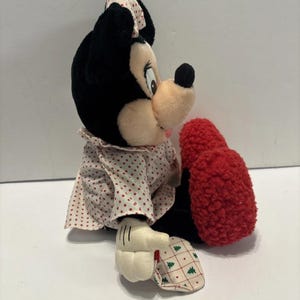 May include: A Minnie Mouse soft toy with black fur, a pink nose, and a red bow with white spots. She wears a white dress with red spots and a red textured foot. Her white gloves have black lines.