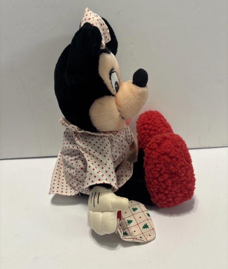 May include: A Minnie Mouse soft toy with black fur, a pink nose, and a red bow with white spots. She wears a white dress with red spots and a red textured foot. Her white gloves have black lines.