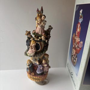 Puede incluir: Una detallada figura con un diseño de castillo con múltiples niveles y figuras. El nivel superior muestra a una pareja abrazándose, mientras que otras figuras están posicionadas alrededor de la estructura. La paleta de colores incluye acentos rosas, marrones y dorados.