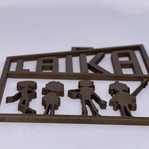 May include: A bronze-coloured metal cutout featuring the word "LAIKA" above the silhouettes of three figures. The figures appear to be children, with one holding a star-shaped object. The cutout is likely a decorative item.