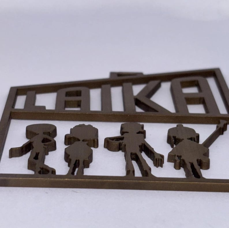 May include: A bronze-coloured metal cutout featuring the word "LAIKA" above the silhouettes of three figures. The figures appear to be children, with one holding a star-shaped object. The cutout is likely a decorative item.