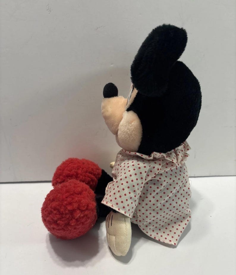 May include: A Minnie Mouse soft toy, with black ears, a black nose, and a cream-coloured face. She is dressed in a white dress with red polka dots and red, textured shoes. A classic Disney character.