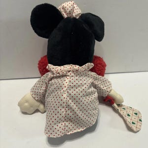 May include: A Minnie Mouse plush toy, seen from behind, dressed in a white and red polka dot dress and bow. The doll has black ears and a red pom-pom tail. It holds a small, patterned fan with green Christmas trees.