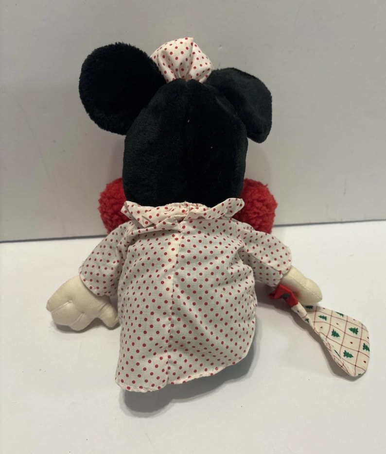 May include: A Minnie Mouse plush toy, seen from behind, dressed in a white and red polka dot dress and bow. The doll has black ears and a red pom-pom tail. It holds a small, patterned fan with green Christmas trees.
