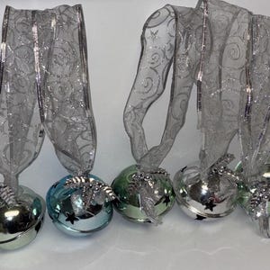 以下が含まれることがあります： 透明なシルバーリボンが付いた、銀色のジングルベルのコレクション。ベルはシルバー、ブルー、グリーンの色合いで、星の切り抜きがあります。各ベルには装飾的なシルバーのリボンが付いており、ホリデーデコレーションに最適です。