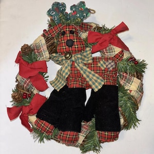 Könnte beinhalten: Ein Weihnachtskranz mit einem karierten Rentier mit schwarzen Stiefeln, roten Schleifen und einer Glocke. Der Kranz besteht aus natürlichen Zweigen und ist mit kariertem Stoff und Tannenzapfen verziert.