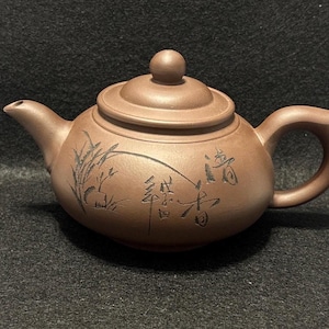 以下が含まれることがあります： 蓋と取っ手が付いた茶色のセラミックティーポット。 ティーポットには注ぎ口があり、黒いカリグラフィーと自然をテーマにしたイラストで装飾されています。 ティーポットはおそらくお茶を淹れるために使用されます。