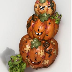 May include: A stacked pumpkin decoration with a smiling face, leaf accents, and a frog figurine. The orange pumpkins have cut-out designs and are topped with green leaves. A green frog sits at the base. A whimsical Halloween decoration.