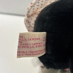 May include: A close-up of a black Minnie Mouse plush toy. The toy has a white bonnet with red polka dots. A tag is visible with text including "Disney Company" and "X'MAS MINNIE FLOPPY".