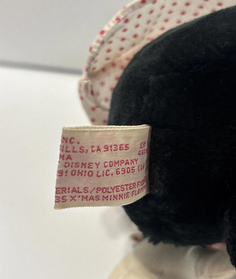 May include: A close-up of a black Minnie Mouse plush toy. The toy has a white bonnet with red polka dots. A tag is visible with text including "Disney Company" and "X'MAS MINNIE FLOPPY".