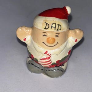 Puede incluir: Una figura de cerámica de un personaje parecido a un muñeco de nieve con un gorro de Papá Noel rojo y la palabra "DAD" en la frente. La figura tiene una cara sonriente, los brazos extendidos y una corbata a rayas rojas y blancas. La figura lleva una camisa blanca con lunares rojos y detalles verdes.