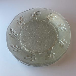 May include: A stack of clear glass plates with a textured surface and a decorative leaf design around the rim. The center of the plates has a dotted pattern. The plates are a light, neutral color.
