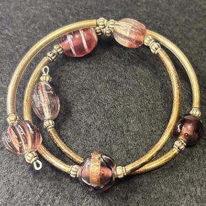 May include: A two-strand bracelet with gold-toned metal bands. The bracelet features various glass beads in shades of pink and purple, with striped and textured designs. The beads are interspersed along the bands, creating a decorative accessory.