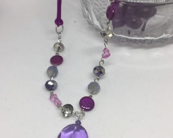 Könnte beinhalten: Eine violette und silberne Perlenkette mit einem großen, runden, violetten Anhänger. Die Kette besteht aus verschiedenen Perlen, darunter runde, facettierte Perlen und flache, runde Perlen.