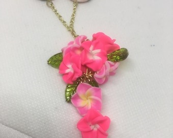Peut inclure: Un collier doré avec un motif floral. Le collier comporte deux pendentifs ronds avec un motif floral. Le pendentif principal est un groupe de fleurs roses avec des feuilles vertes. Le collier est sur une surface blanche.