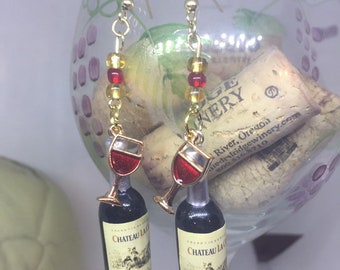 May include: A pair of gold dangle earrings with red and gold glass beads. Each earring features a miniature wine bottle with a red wine glass on top. The bottle label reads "Chateau La... Cabernet Sauvignon".