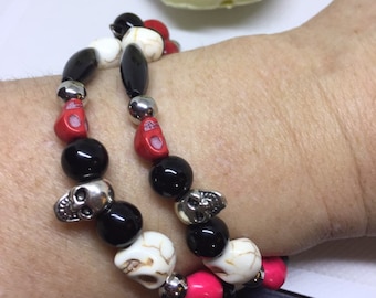May include: Two bracelets with black, white, and red beads. The beads are skull-shaped and some are round. The bracelets are on a person's wrist.