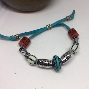 Pode incluir: Um bracelete de couro turquesa com contas prateadas e laranja. O bracelete tem uma conta turquesa com detalhes prateados, duas contas brancas com listras pretas e duas contas laranja com detalhes prateados.