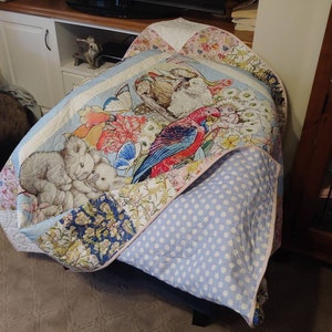 May include: A vibrant quilt with a parrot, koalas, butterflies, and floral designs. It has a light blue border and a light blue polka dot reverse. The quilt is draped over a chair.