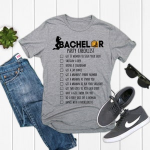 May include: A gray t-shirt with a black graphic of a checklist for a bachelor party. The checklist includes items such as "Get 10 women to sign your shirt", "Shotgun a beer", and "Dance with a bachelorette".