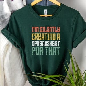 Puede incluir: Una camiseta verde oscuro con el texto "I'm silently creating a spreadsheet for that" en una fuente de estilo retro con colores naranja, amarillo y verde.