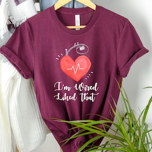 Puede incluir: Camiseta color burdeos con un gráfico blanco de un corazón con una línea de latido del corazón y un dispositivo médico. El texto "I'm Wired Like That" está impreso debajo del corazón.