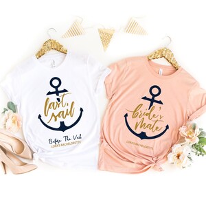 Może przedstawiać: Dwie koszulki w kolorze białym i brzoskwiniowym z grafiką kotwicy i tekstem "last sail" i "bride's mate" złotymi literami. Koszulki wiszą na złotych wieszakach i mają nadrukowany tekst "Before The Veil Lora's Bachelorette" na dole.