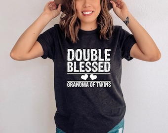 Może przedstawiać: Ciemnoszara koszulka z białym napisem "DOUBLE BLESSED GRANDMA OF TWINS". Koszulka ma dwa białe serca pod napisem "DOUBLE BLESSED". Osoba na zdjęciu ma na sobie brązowy kapelusz i niebieskie dżinsy.
