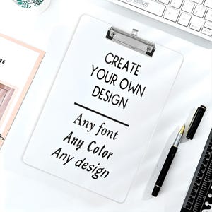 May include: A white clipboard with a silver metal clip at the top. The surface features black text that reads: CREATE YOUR OWN DESIGN, Any font, Any Color, Any design. The clipboard is positioned on a white surface next to a black pen.