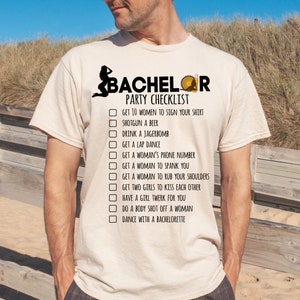 May include: A beige t-shirt with a checklist for a bachelor party. The checklist includes items like "Get 10 women to sign your shirt" and "Dance with a bachelorette".