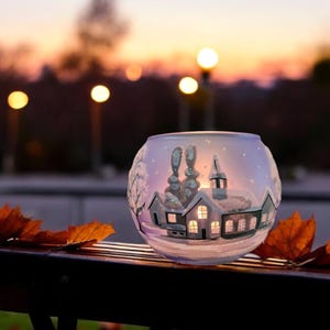 Könnte beinhalten: Ein Glas-Kerzenhalter mit einer darauf gemalten Winterdorfszene. Der Kerzenhalter steht auf einer Holzbank mit verstreuten Herbstblättern. Der Hintergrund ist ein verschwommener Sonnenuntergang mit Straßenlaternen.