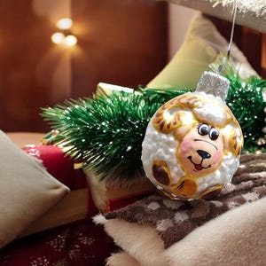 May include: A white Christmas ornament shaped like a sheep with gold accents and a smiling face. The ornament is hanging from a branch of a Christmas tree.