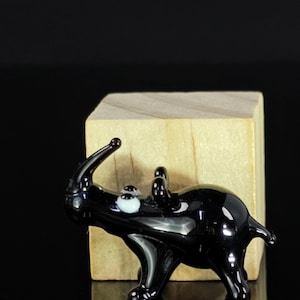 May include: A small black glass rhinoceros figurine with white eyes. The figurine is standing on a wooden block.