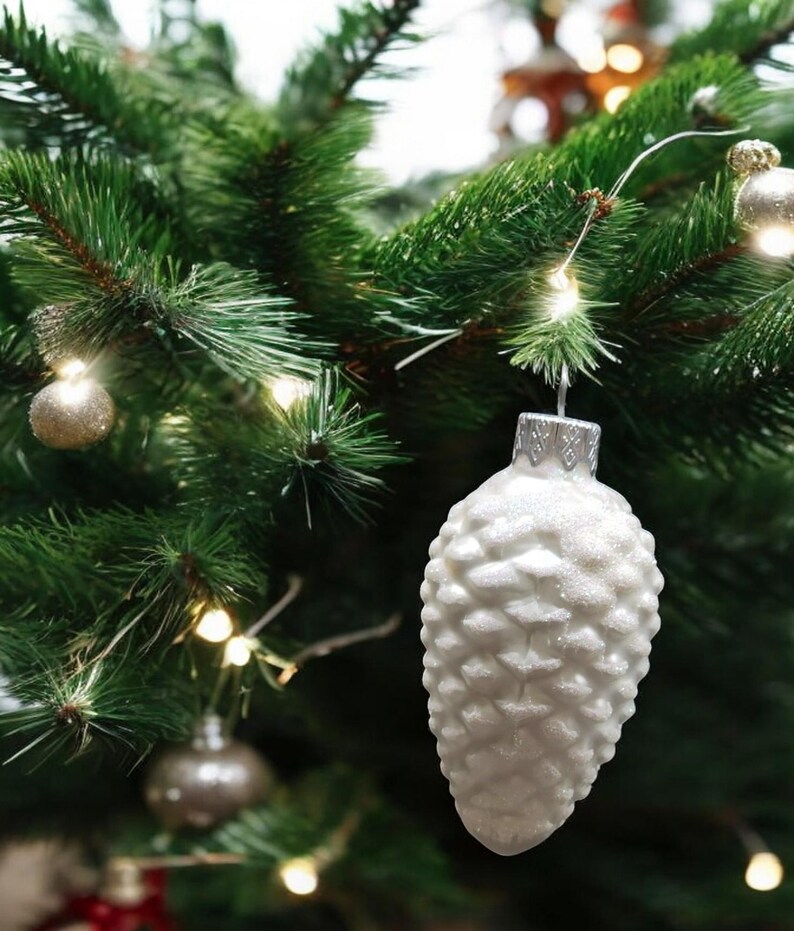 May include: White pine cone shaped Christmas ornament with a glittery finish hanging on a green Christmas tree.