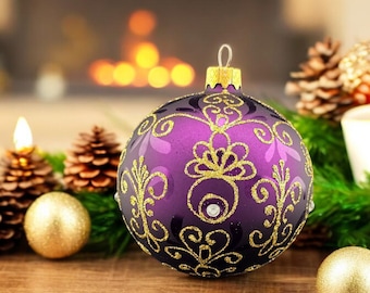 May include: A purple Christmas ornament with gold filigree design and a gold cap. The ornament is surrounded by pine cones, gold baubles, and greenery, creating a festive holiday scene. The background features a blurred fireplace.