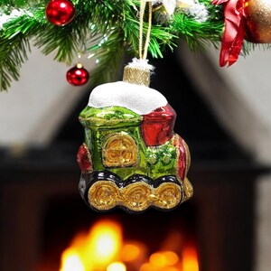 May include: A green and red glass Christmas ornament shaped like a train with gold wheels. The ornament has a white snowy top and is hanging from a Christmas tree.