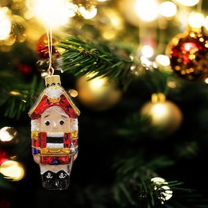 May include: A glass Christmas ornament shaped like a nutcracker soldier. The ornament is red, white, and blue with gold accents. The nutcracker is holding a drum and has a mustache.