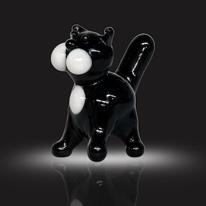 Könnte beinhalten: Eine glänzende schwarze Glasfigur einer Katze mit einem glatten, abgerundeten Körper. Die Katze hat zwei markante weiße, kreisförmige Wangen, einen kleinen weißen Fleck auf der Brust und einen hohen, aufrechten Schwanz. Die Oberfläche ist reflektierend.