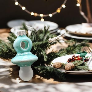 May include: A light blue glass ornament shaped like a baby's pacifier with white polka dots. The ornament has a clear opening in the center and a white frosted base.