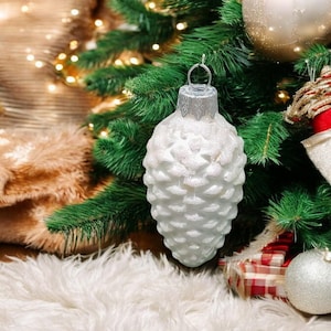 May include: A white, frosted pine cone-shaped Christmas ornament with a silver top hangs on a green Christmas tree.